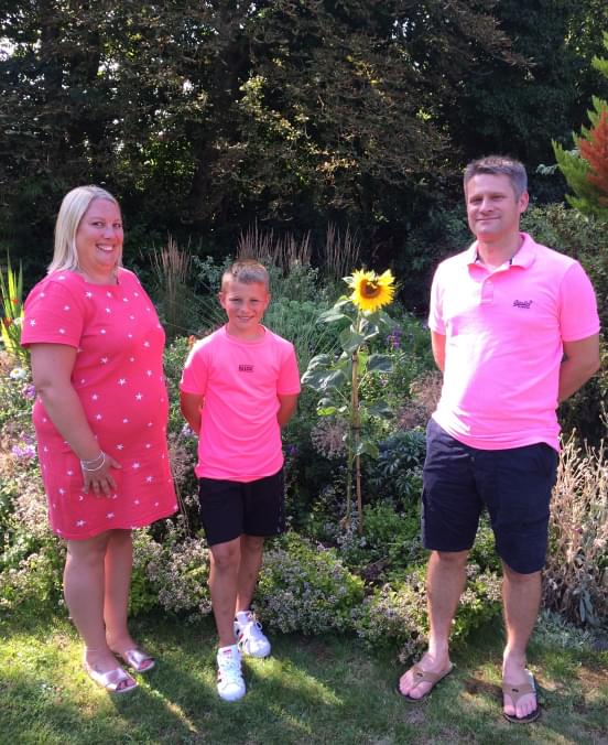 Mum and dad stand either side of their son. They are photographed with a sunflower in a garden.